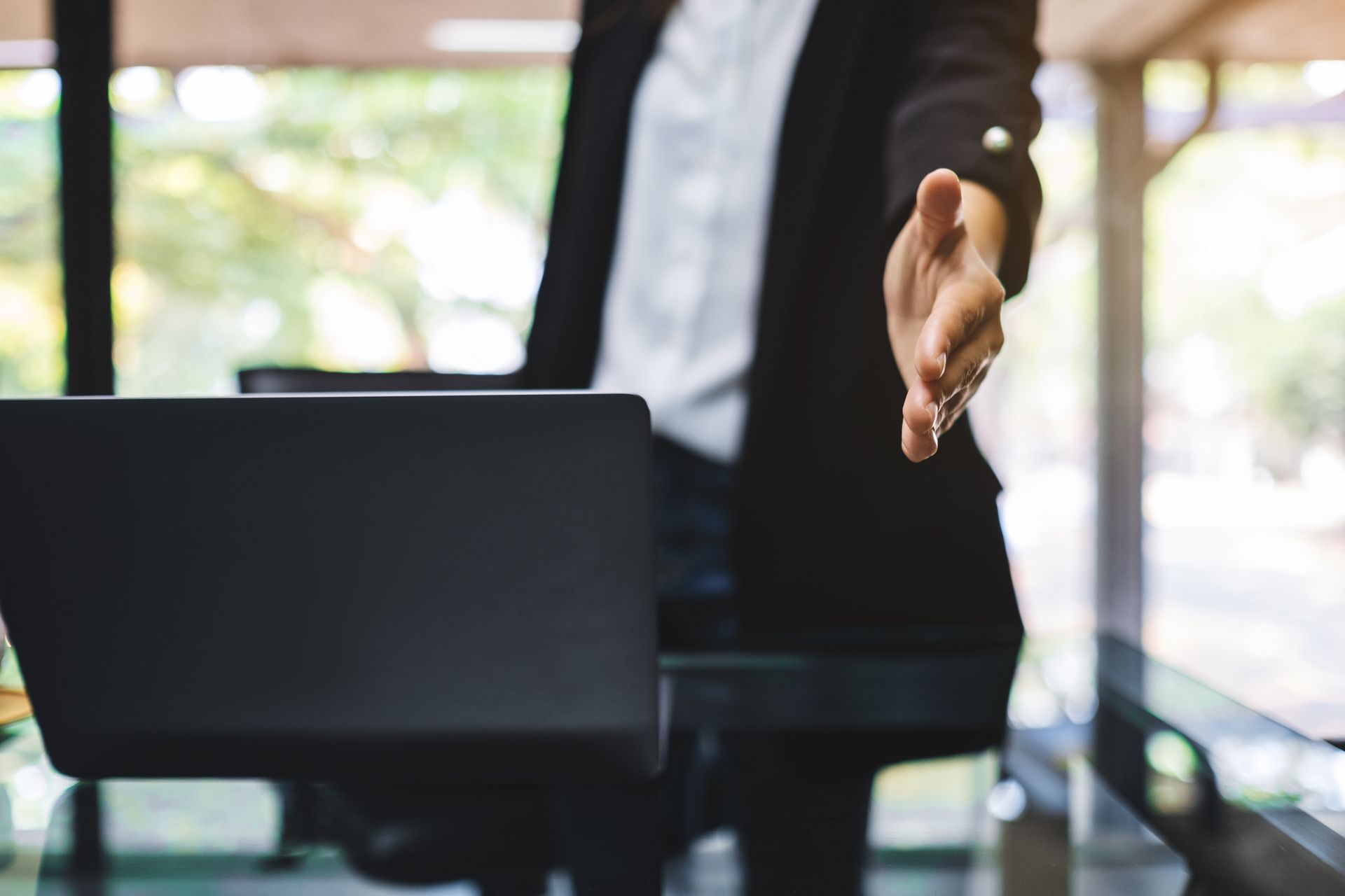 A man is sitting at a desk with a laptop and reaching out to shake someone 's hand.
