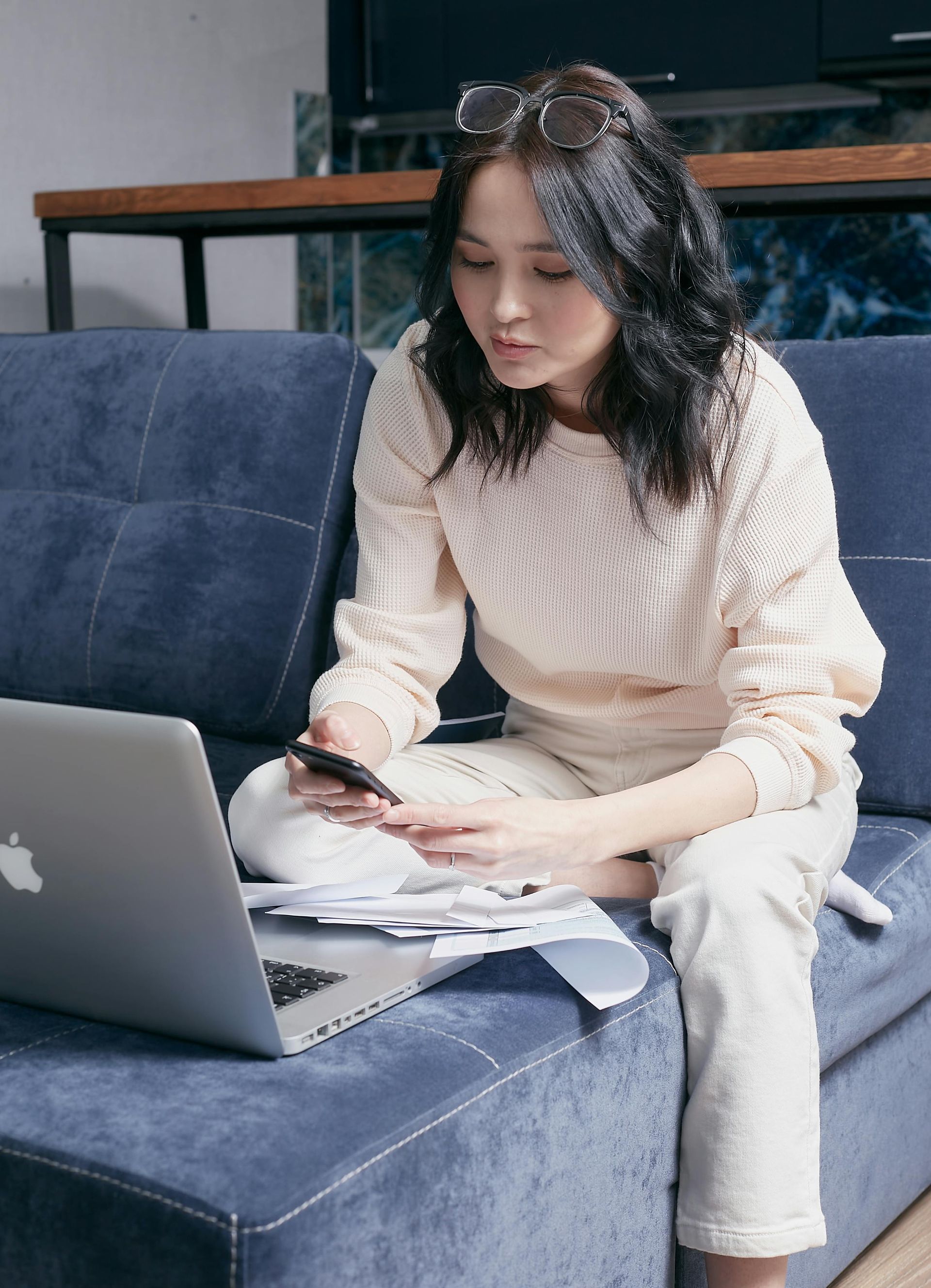 A woman is sitting on a couch using a laptop and a cell phone.