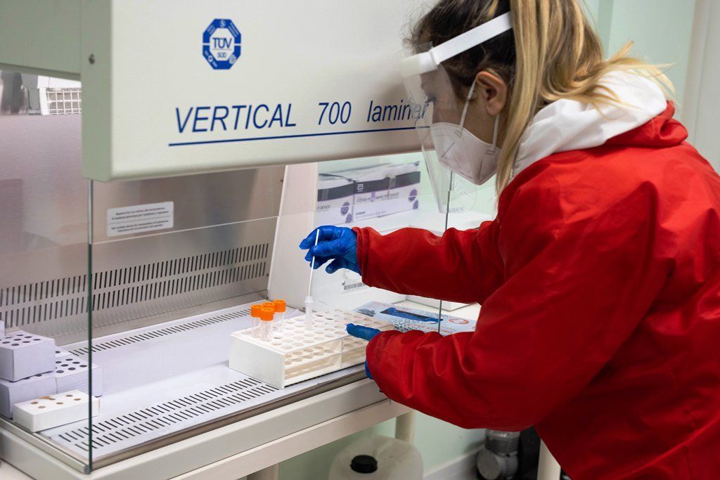 Una persona con il camice rosso lavora con delle provette all'interno di una cappa da laboratorio bianca.