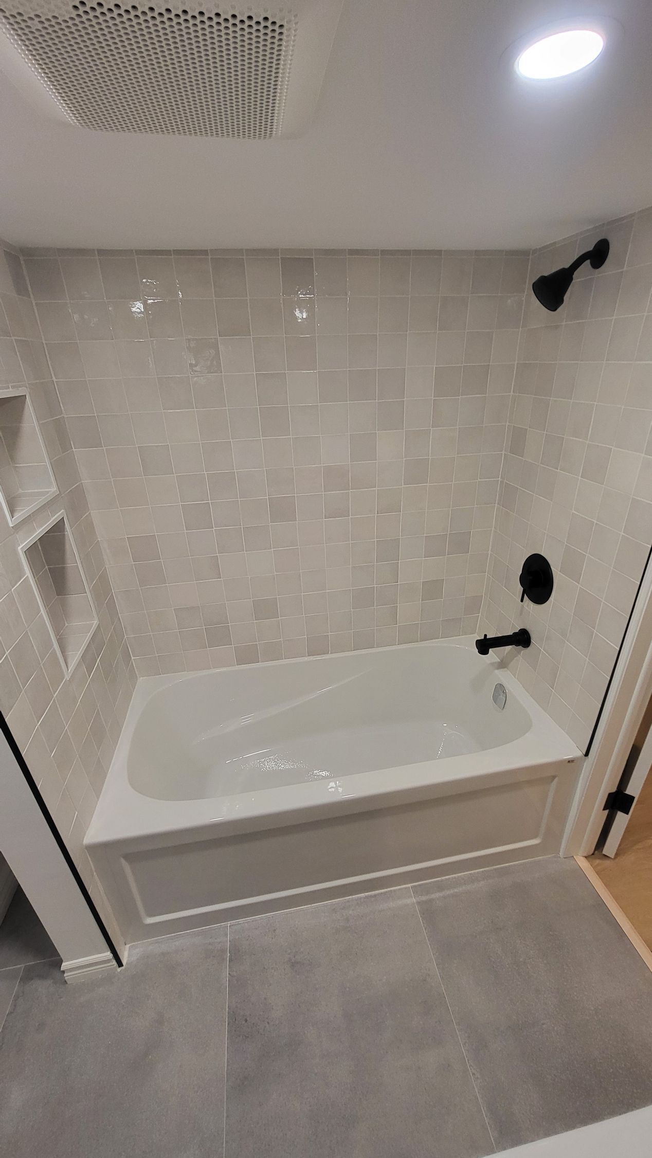 Bathroom with gray tile walls, white bathtub, black fixtures, and recessed shelves.