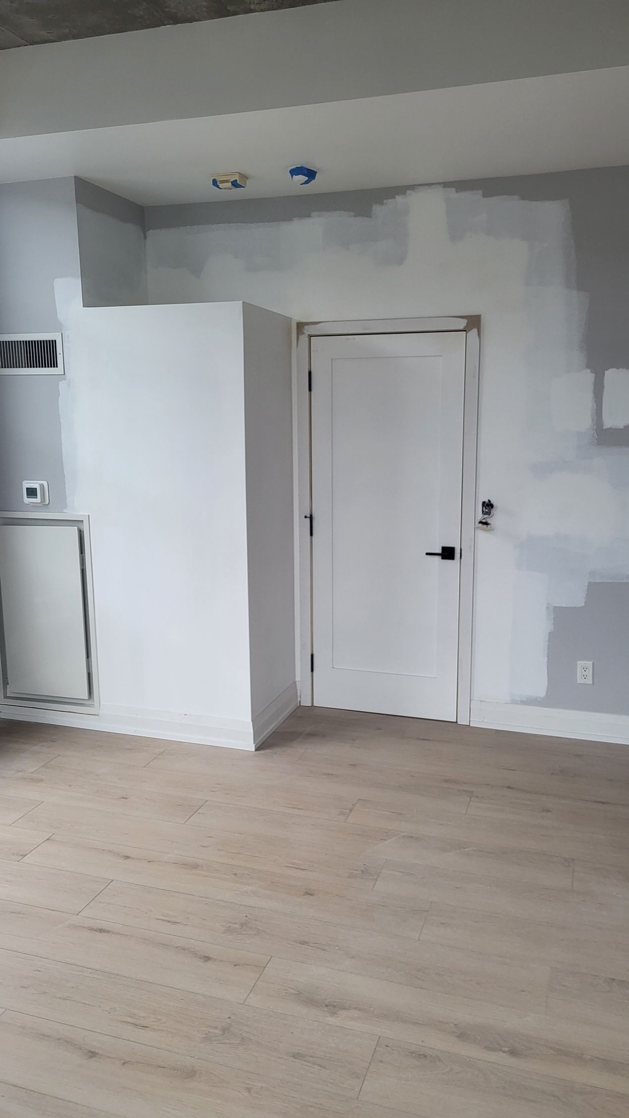 Room with light-colored hardwood floors, white walls, and a white door with black handle. Some gray paint is visible.