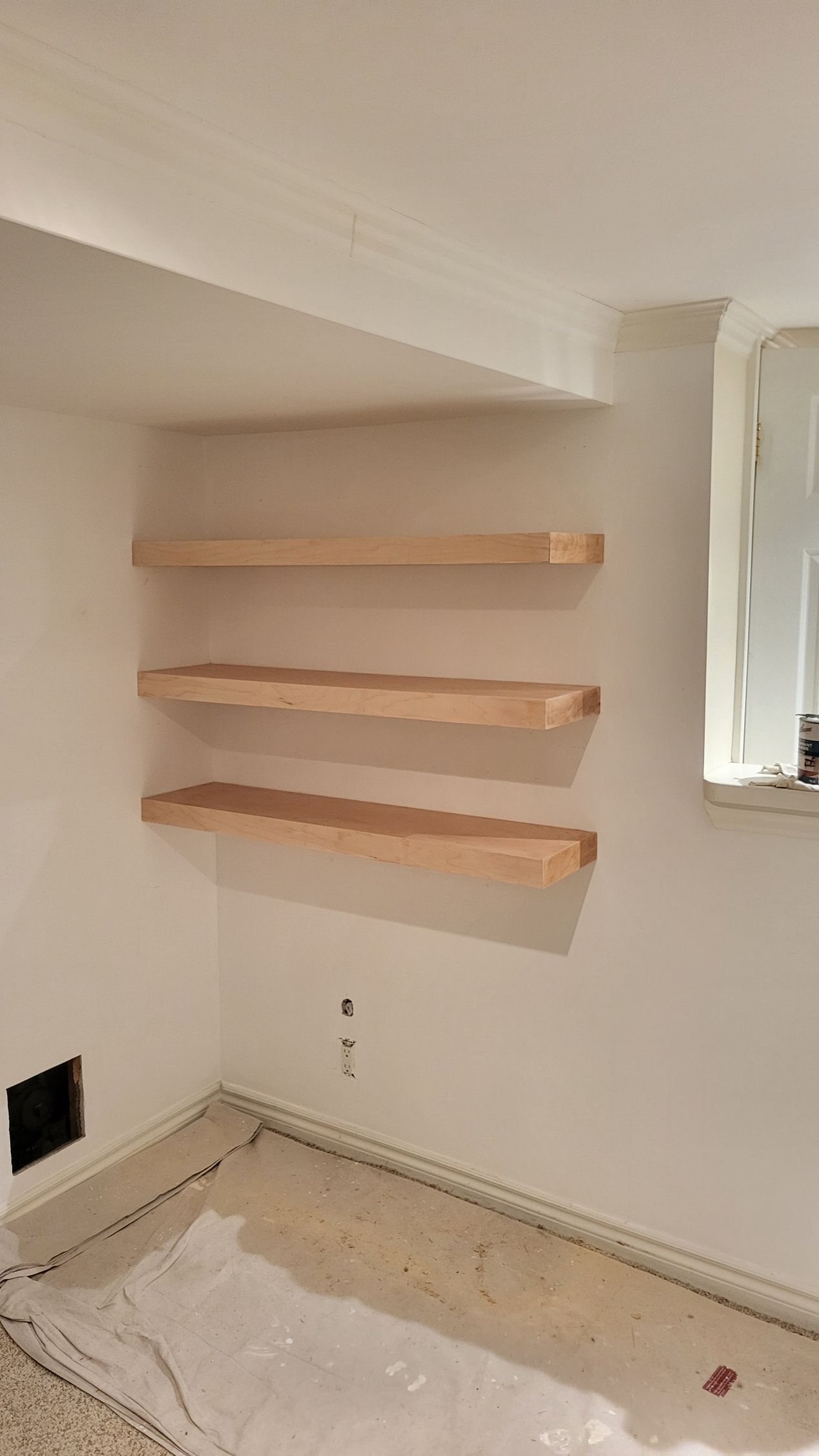Three unfinished wooden shelves on a white wall in a corner.
