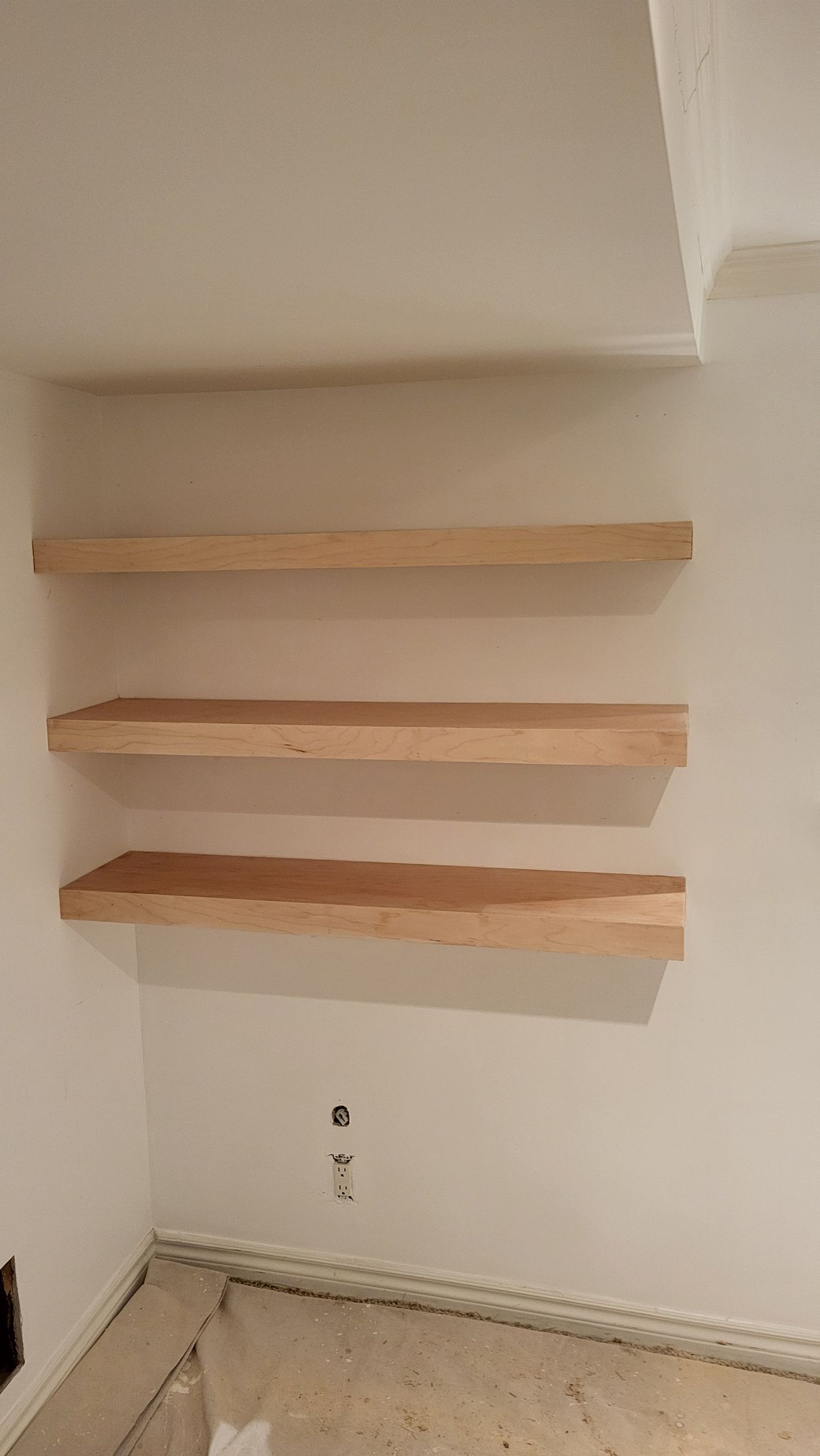 Three unfinished wooden shelves mounted on a white wall in a corner.