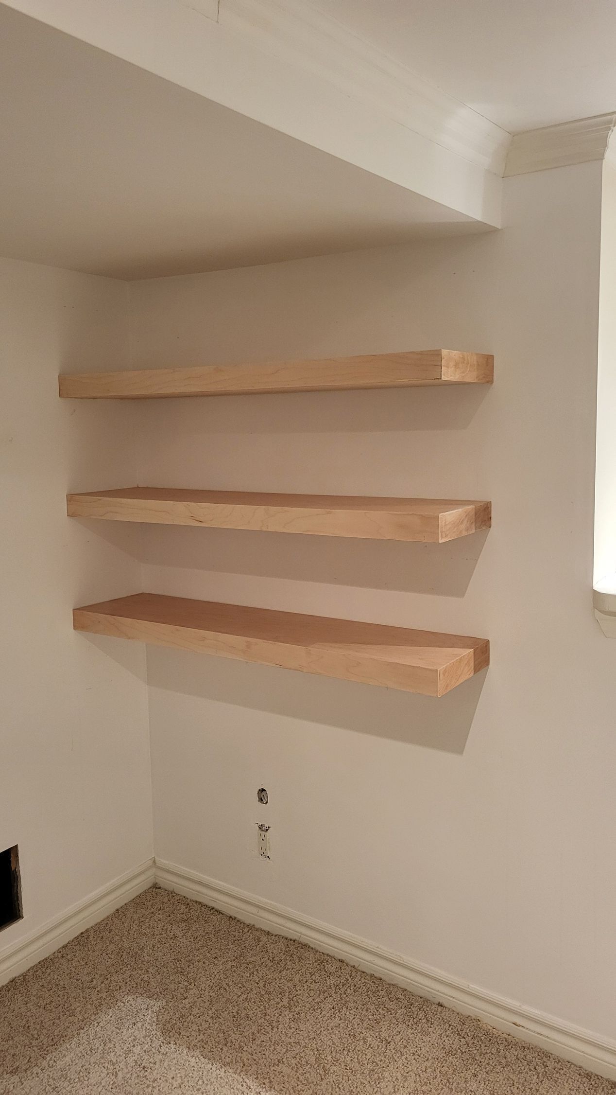 Three unfinished wood shelves mounted on a white wall in a corner of a room with carpet.