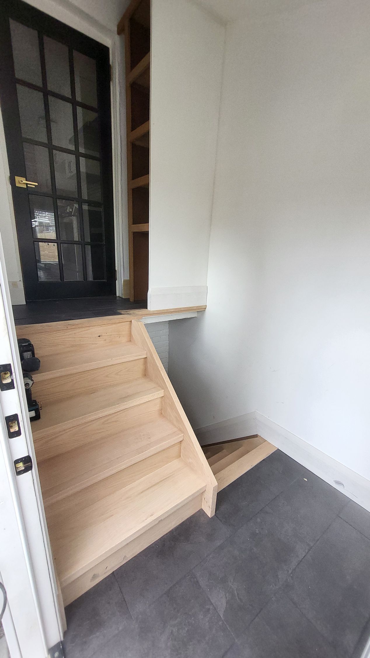Wooden stairs lead up to a black door with glass panes. White walls and built-in shelving are visible.