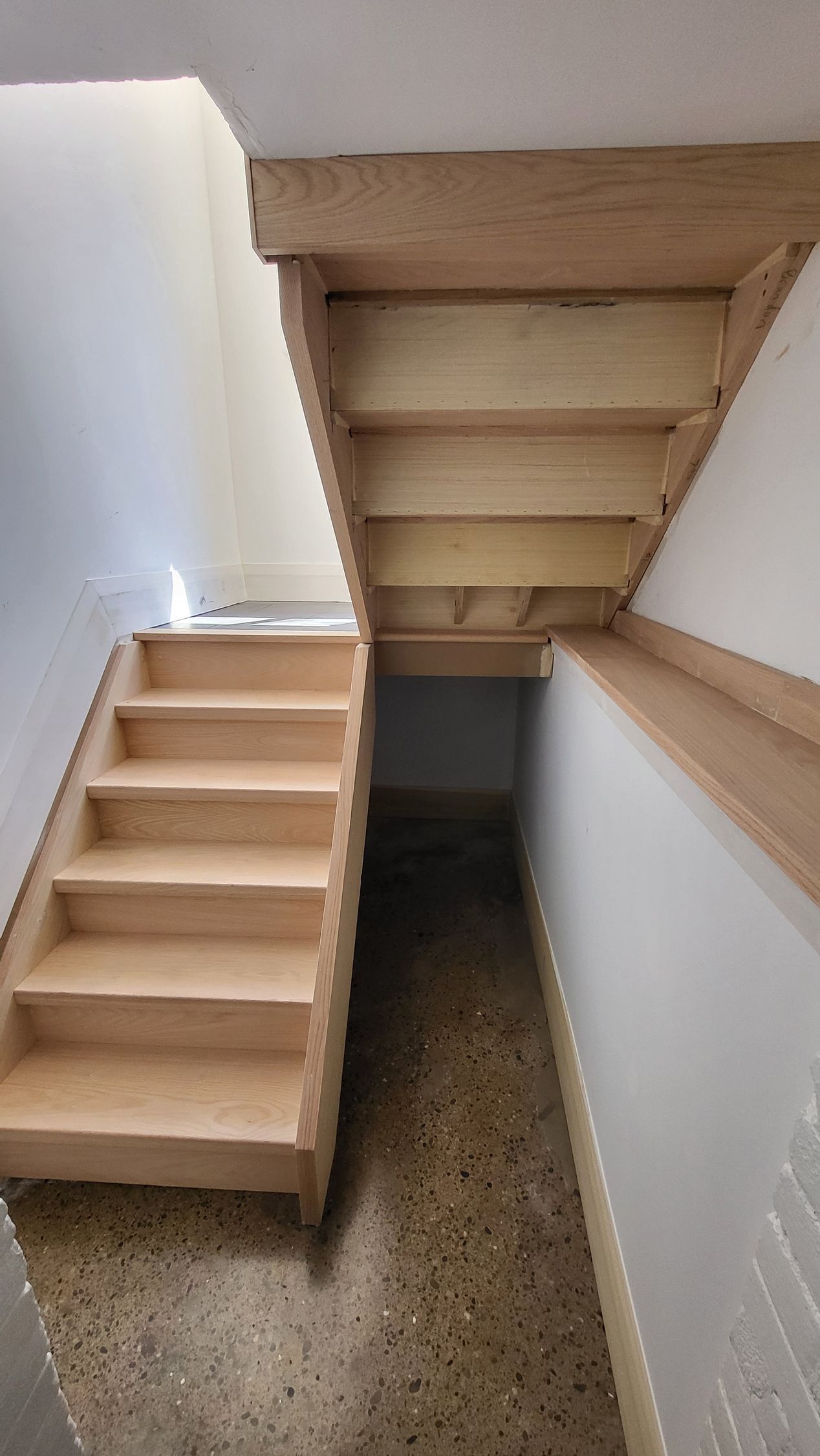 Staircase with wooden steps and railing leading to a lower level, with white walls and a concrete floor.