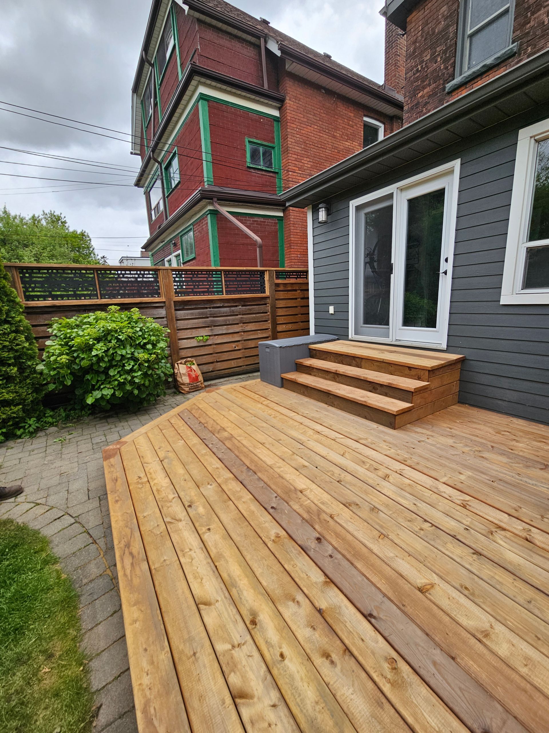 Wooden deck with steps leading to a gray house with a sliding door, next to a brown fence and brick building.