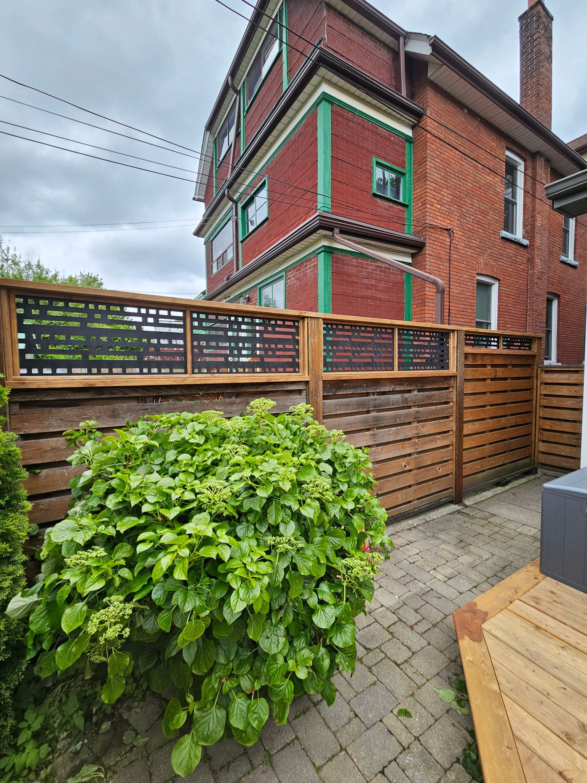 Red brick building with green trim, a wooden fence, and a green bush in a paved yard.