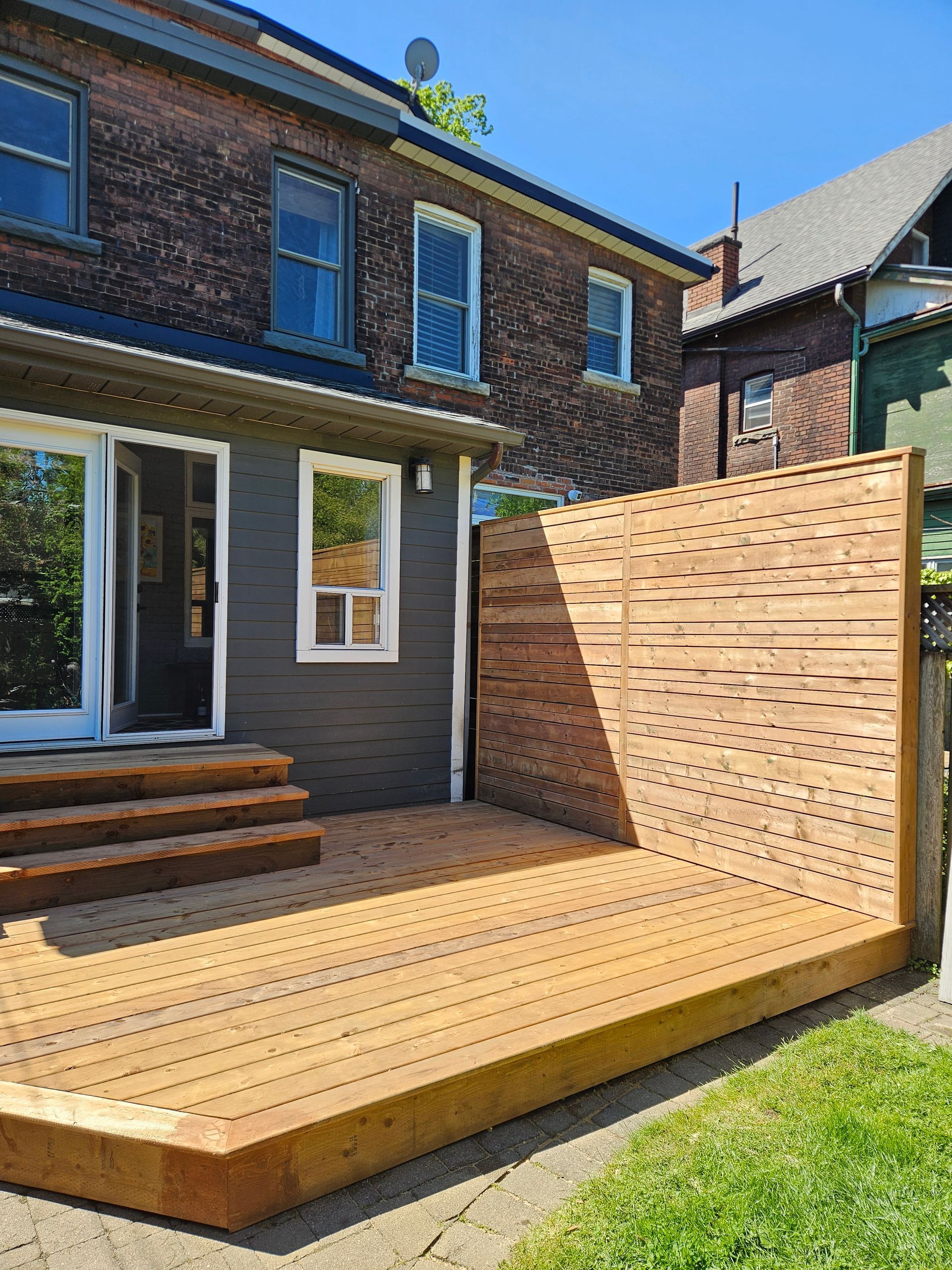 Wooden deck with privacy screen next to a brick building. Bright sunlight, grass in foreground.
