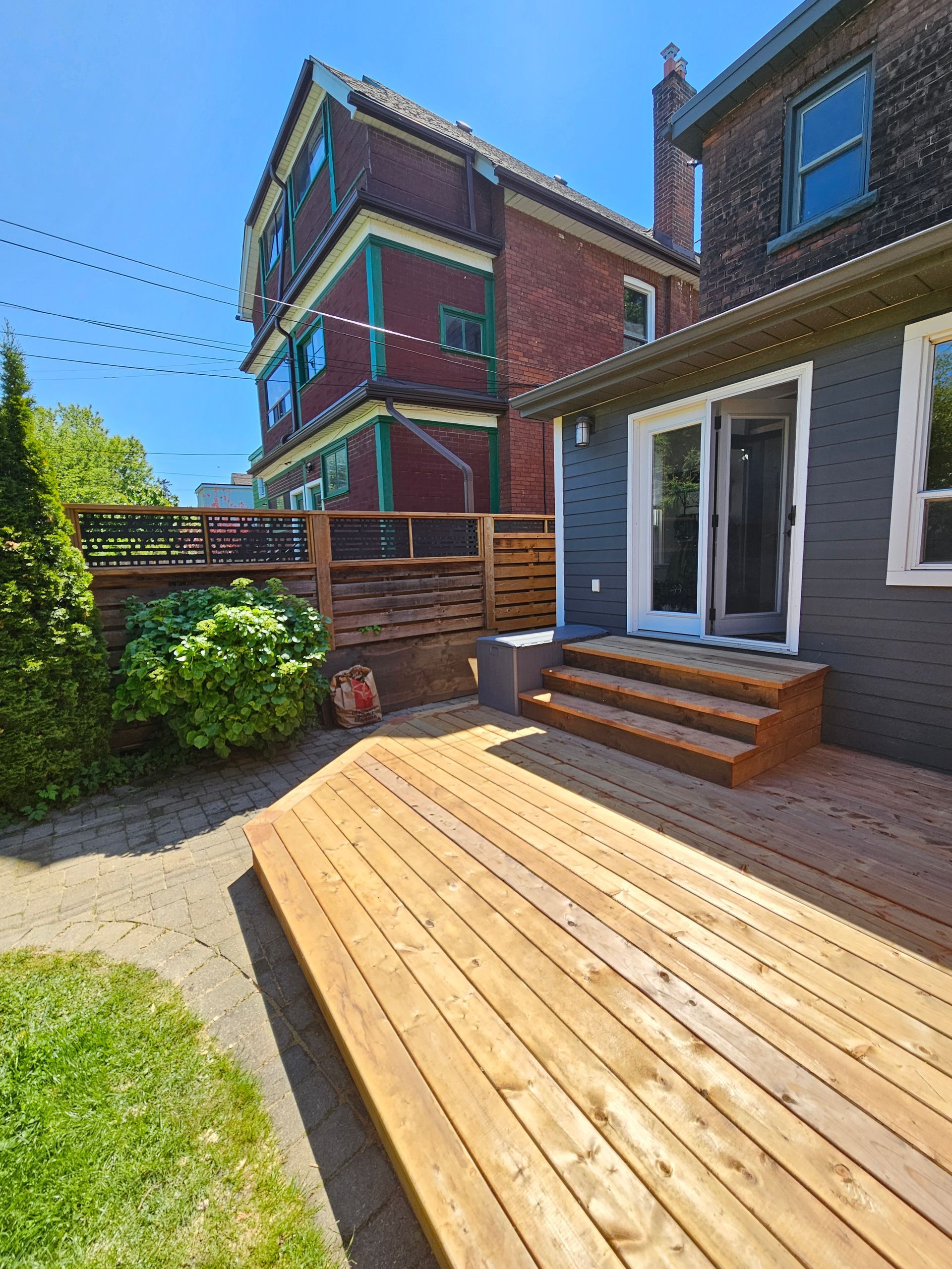 Wooden deck and steps leading to a gray house, with a multi-story brick building in the background.