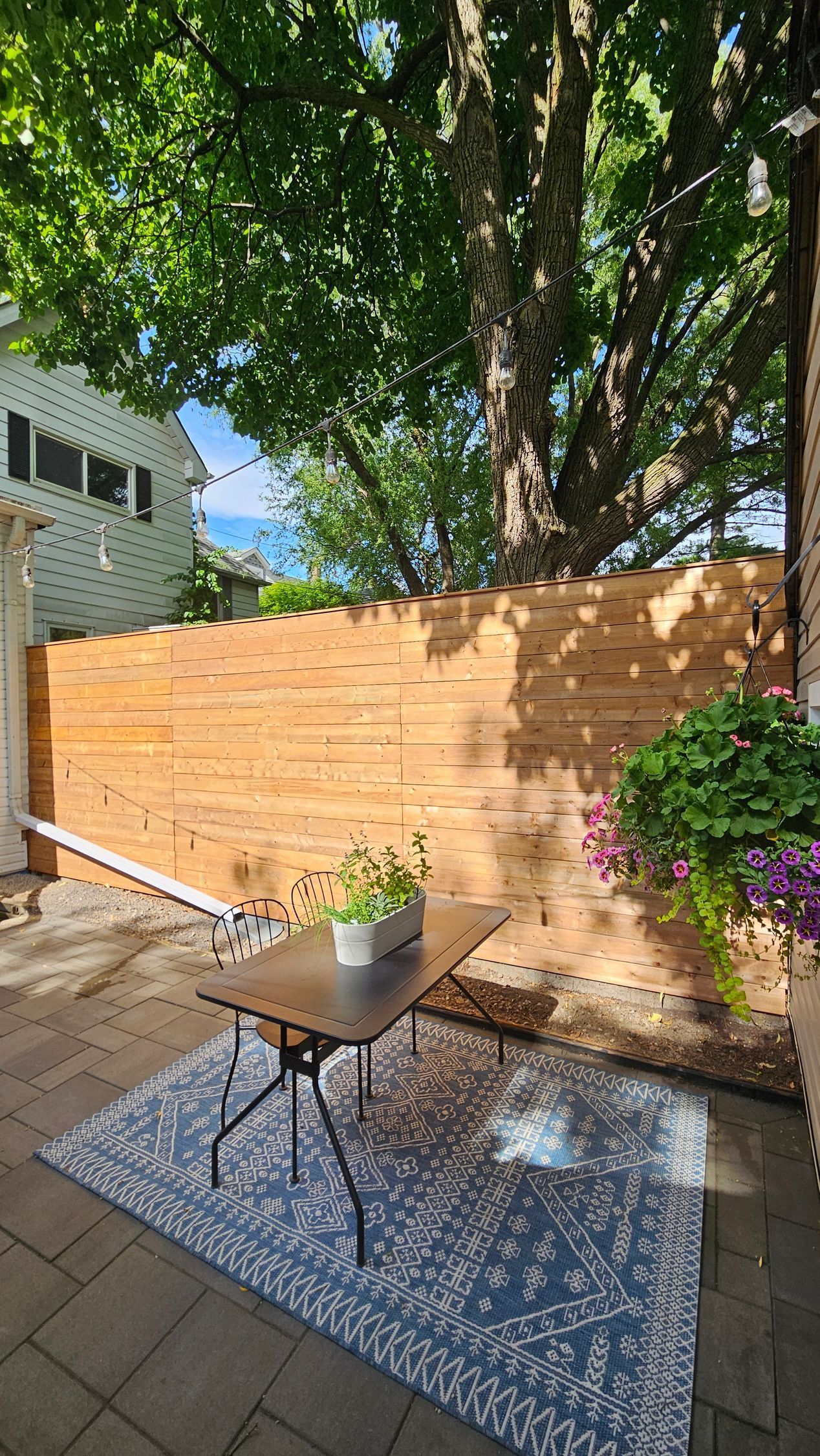 Patio with table, rug, and wooden fence, under tree canopy.