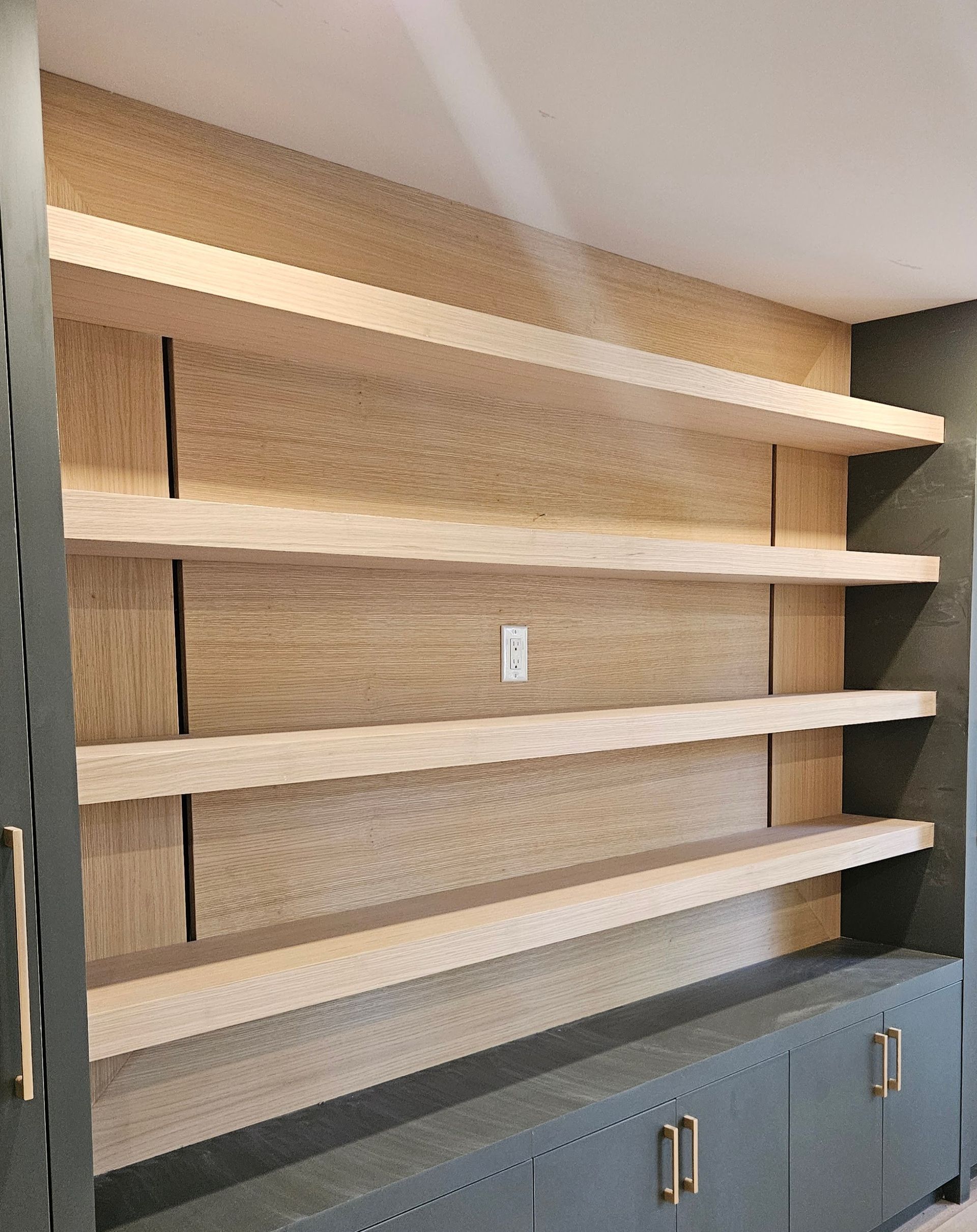 Built-in shelves with light wood shelves and back paneling, over a gray cabinet with brass handles.