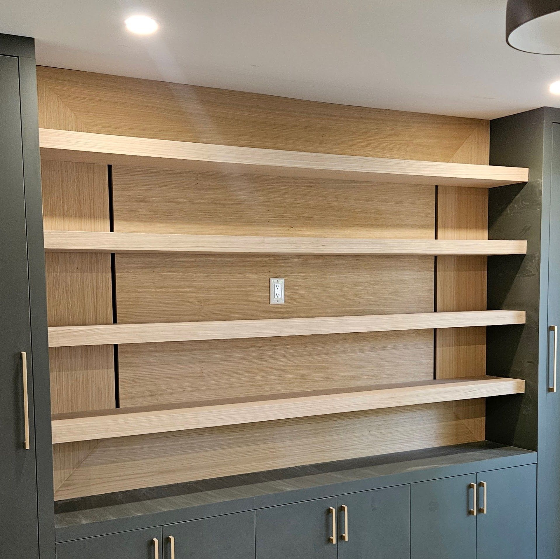 Built-in shelves with light wood shelves and backing, above gray cabinets with gold hardware.