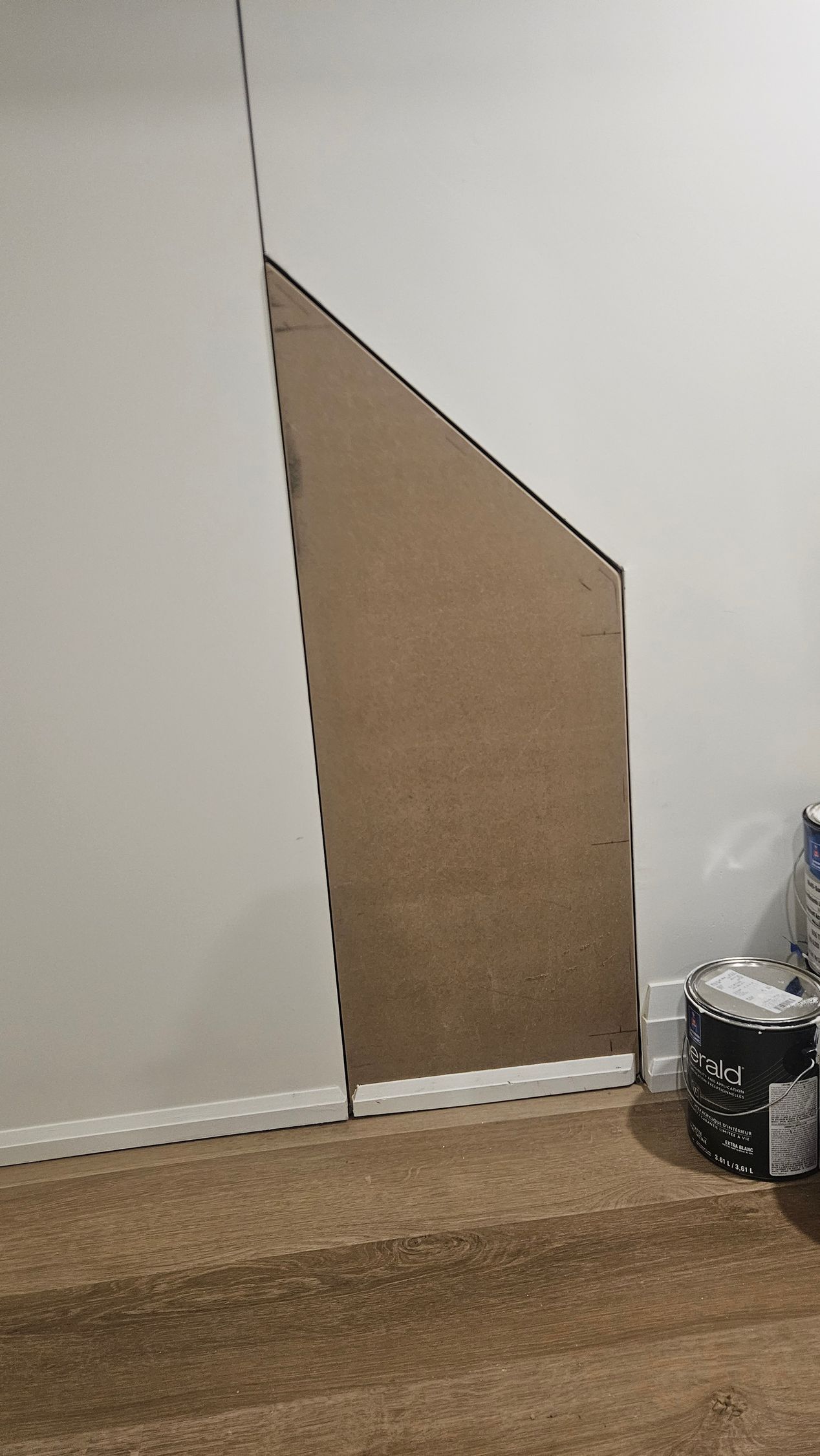A cardboard panel leaning against a white wall with an angled cut, next to a staircase and paint cans.