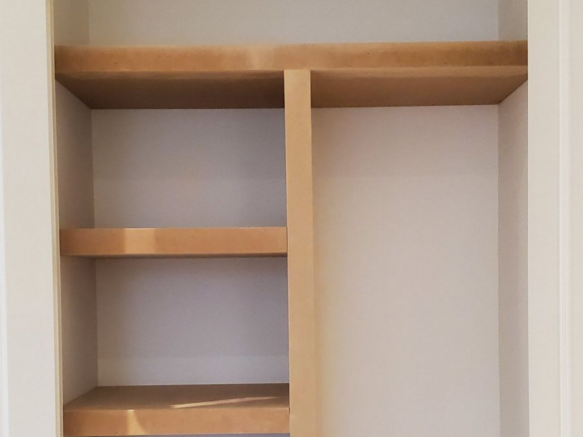 Empty closet with three shelves and a vertical divider. Shelves and divider are tan; walls are white.