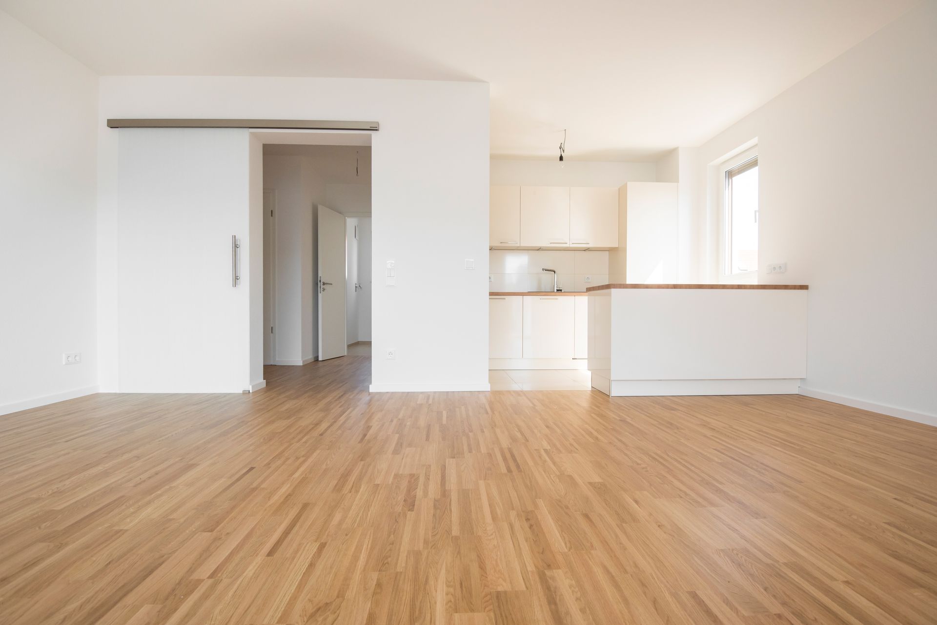 Empty apartment interior with light wood floors, white walls, and a kitchen in the background.