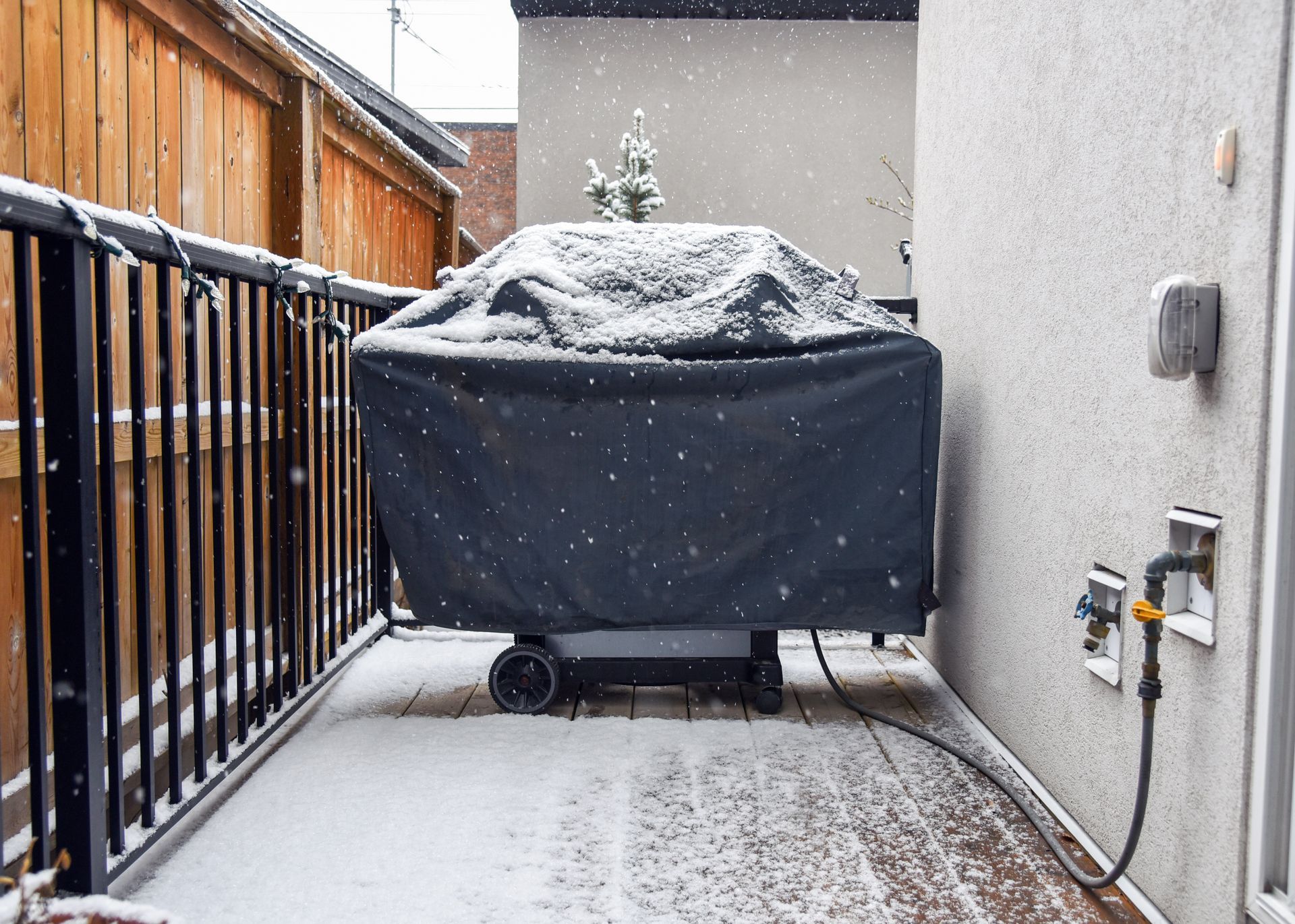 Snowy backyard scene: covered grill on a deck, next to a house wall, with a fence on the left.