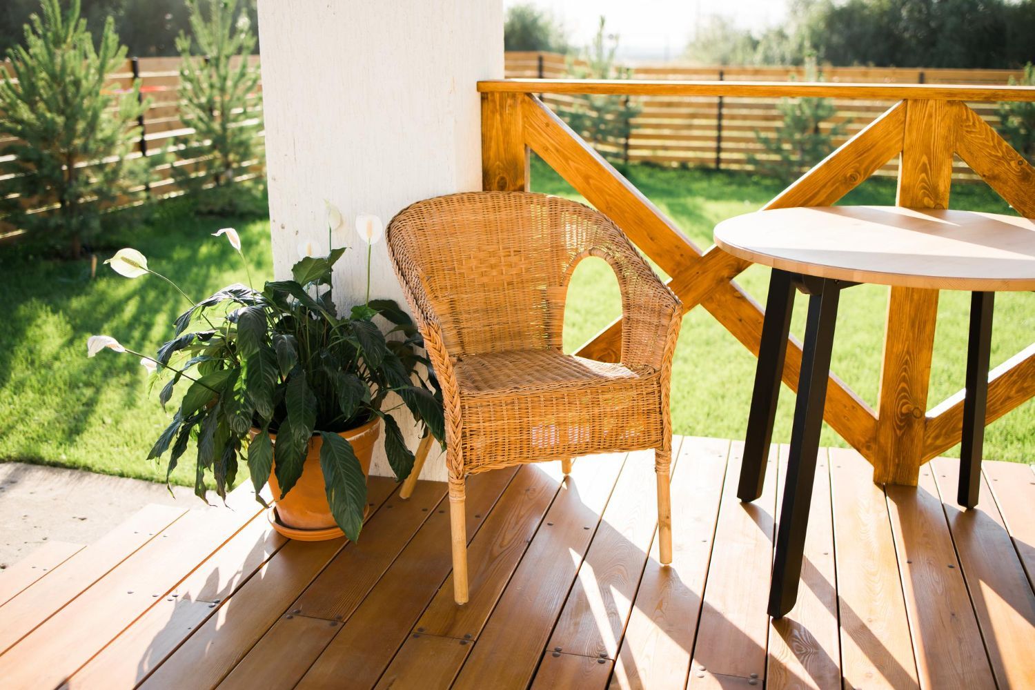 Wicker chair, small round table on a wooden deck, potted plant, wooden fence, sunny outdoor setting.
