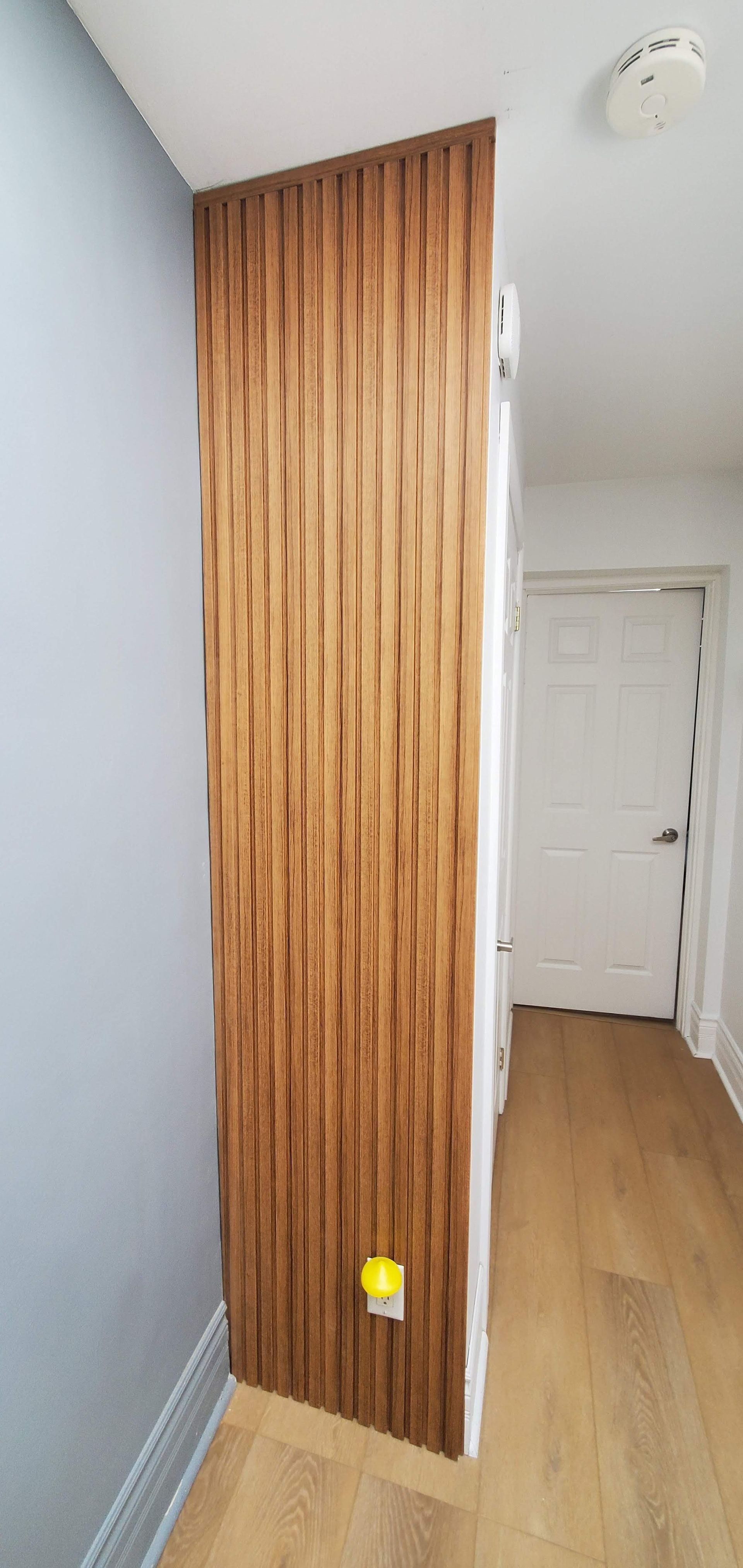 Wooden vertical slat wall panel in a hallway. Blue wall on the left, white door in the distance, and wood floor.