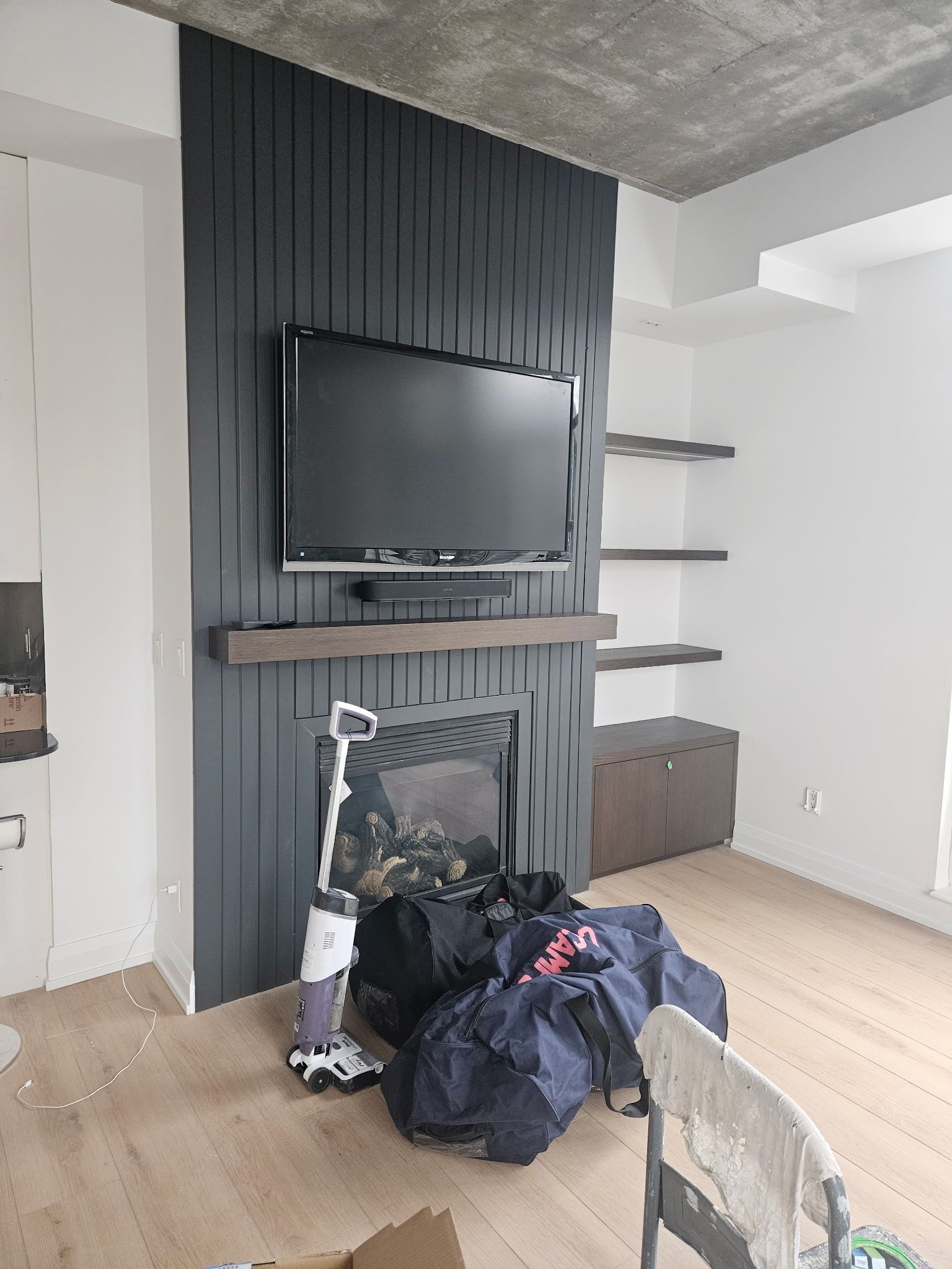 Black accent wall with fireplace and TV, wood shelves, and light wood flooring. Bags and chair in front.