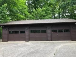 Three-bay brown garage with green doors, set against dense trees and a gravel driveway.