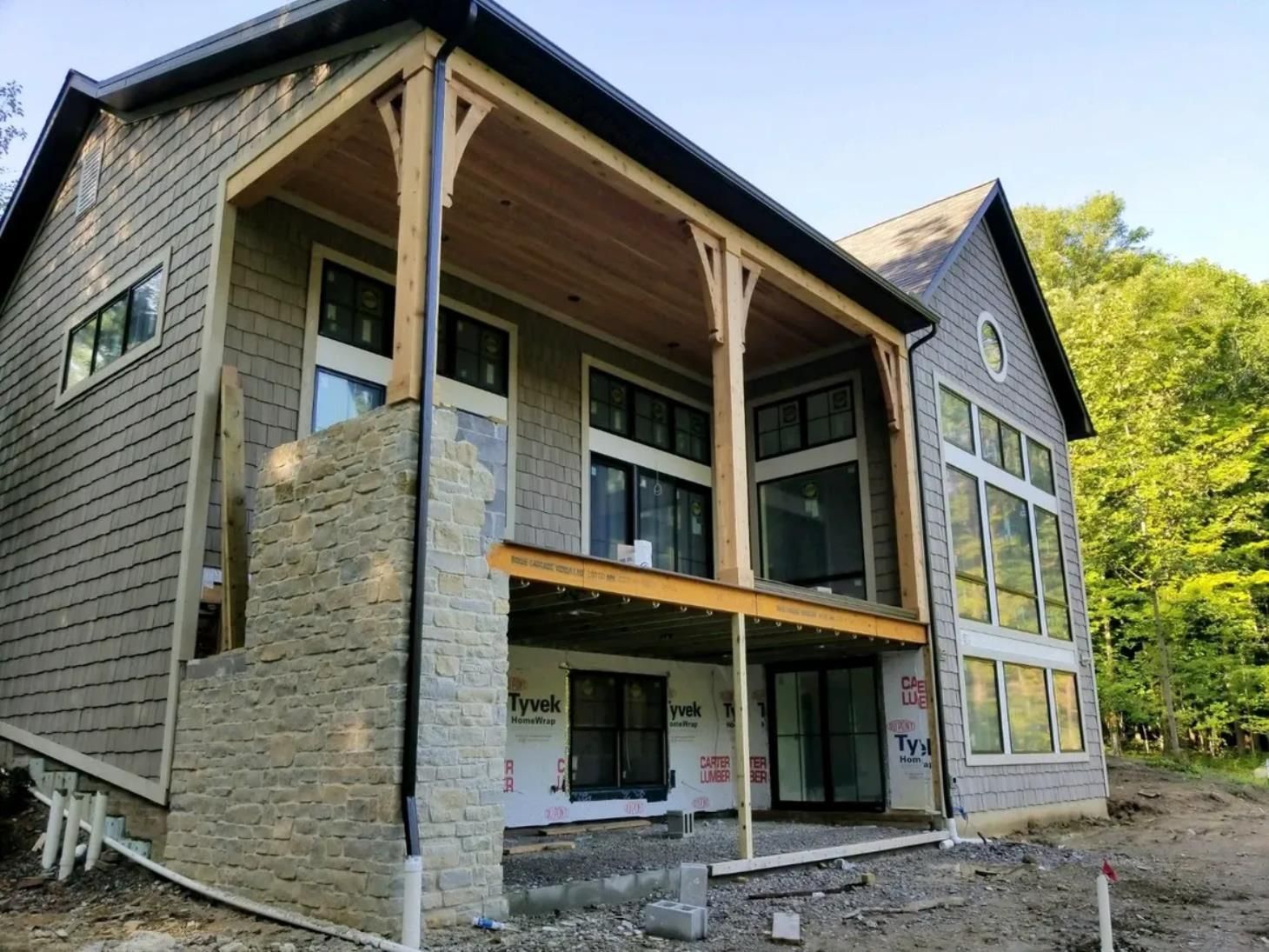 Partially finished house with stone siding, exposed wood framing, and large windows on a wooded lot