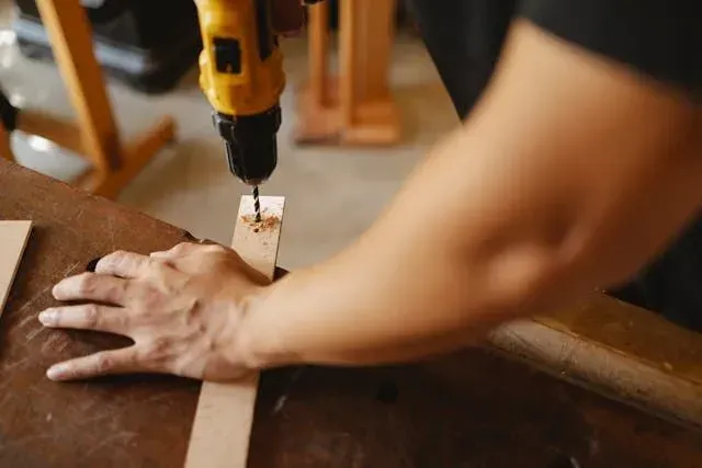 A person using a yellow power drill to make a hole in a thin strip of wood on a workbench.