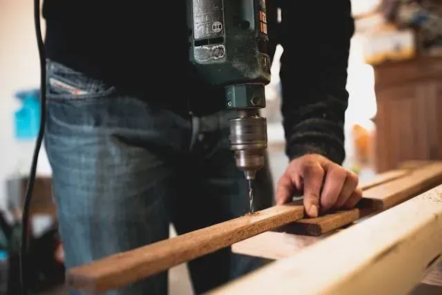 A person using a corded power drill to make a hole in a narrow wooden board in a workshop.