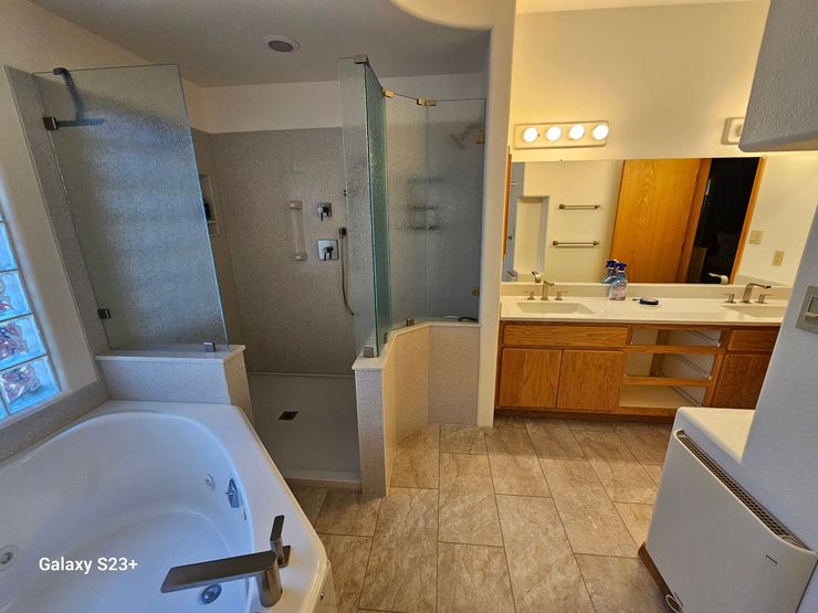 Bathroom with bathtub, glass shower enclosure, double vanity, and wood cabinet under bright vanity lights.ogo
