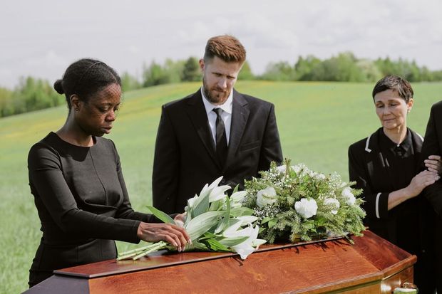 People in formal black attire stand in a grassy field, somberly placing flowers on a casket during a funeral service.