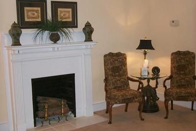 A white fireplace mantel with decor sits beside two patterned armchairs and a small lamp table against a beige wall.