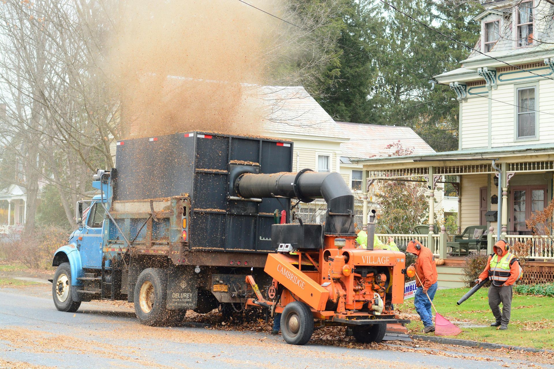 Blue truck with leaf vacuum, workers blowing leaves, residential street.