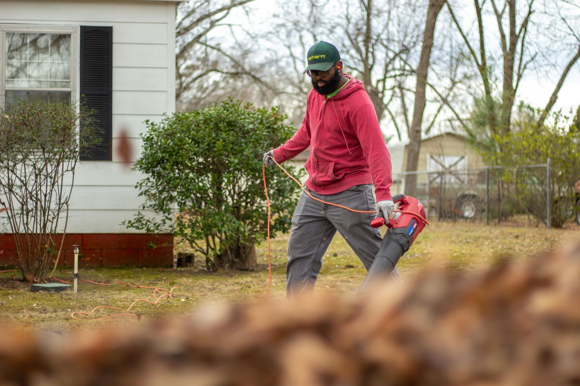 A man in red hoodie uses a leaf blower on a lawn near a house.