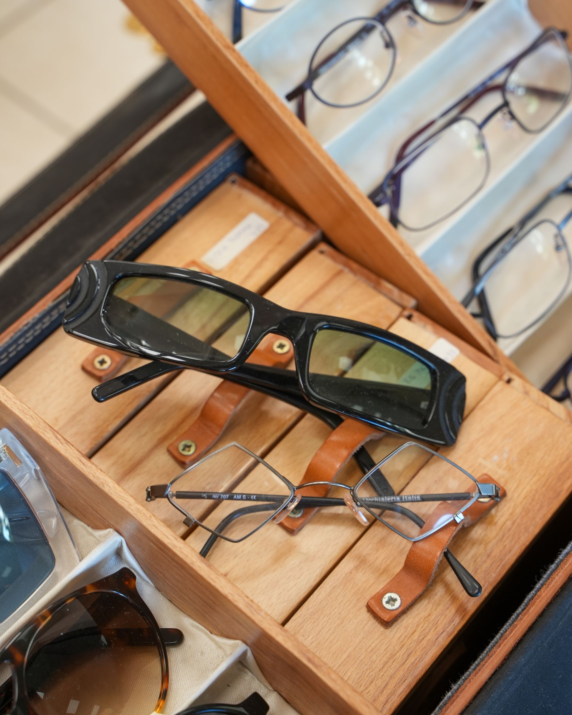 Occhiali esposti in una teca di legno. Sono visibili montature nere e argento, insieme a custodie in pelle marrone.