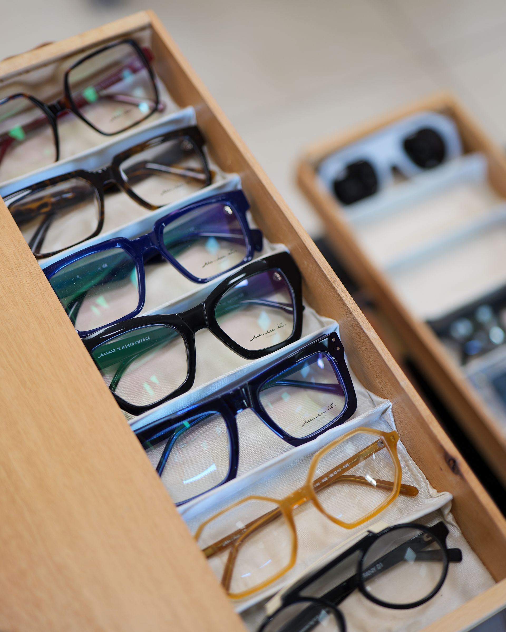 Occhiali esposti in un cassetto di legno. Le montature sono di vari colori e stili, tra cui nero, blu e marrone.