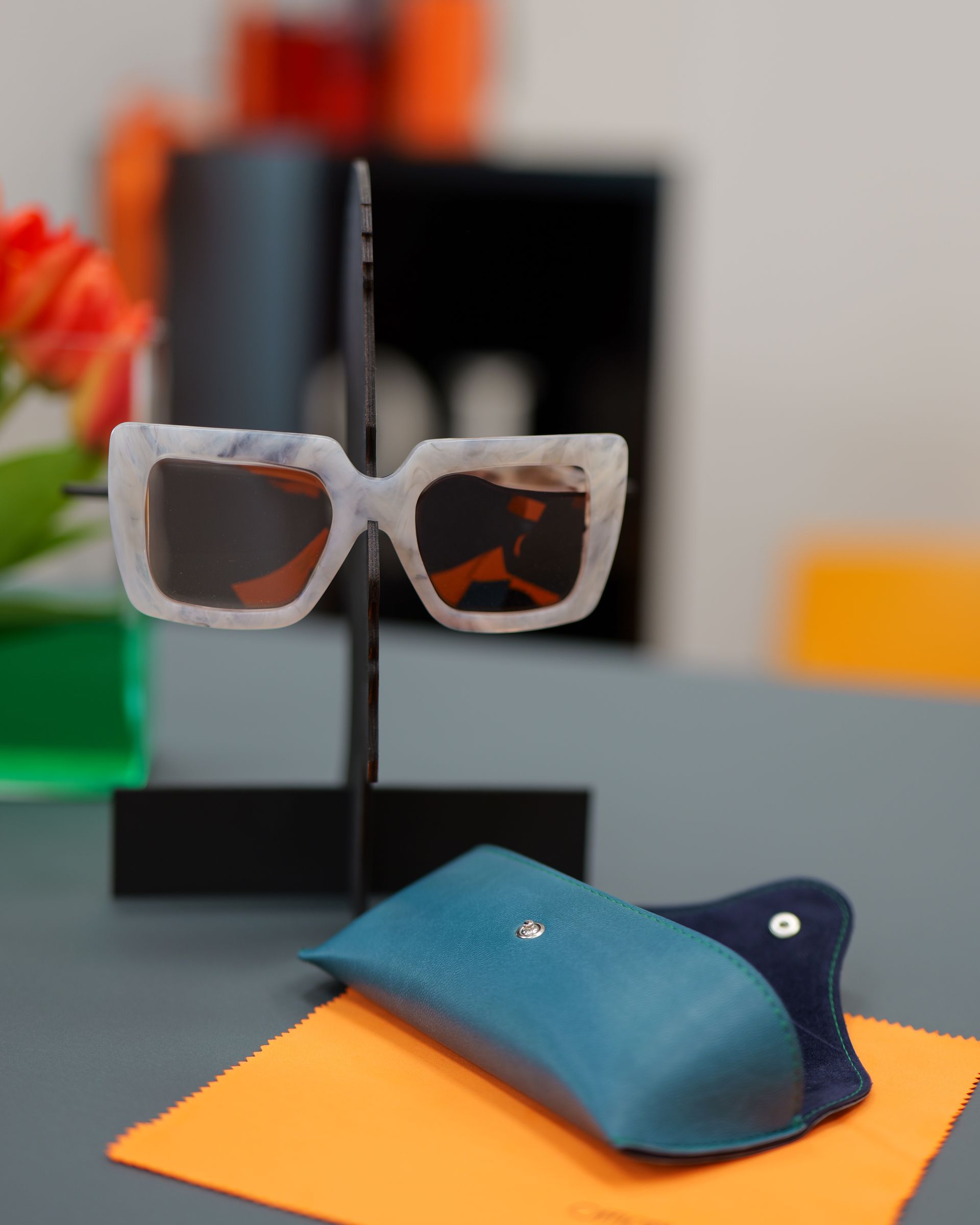 Occhiali da sole con montatura effetto marmo su un supporto nero, accanto a una custodia verde acqua su un panno arancione.
