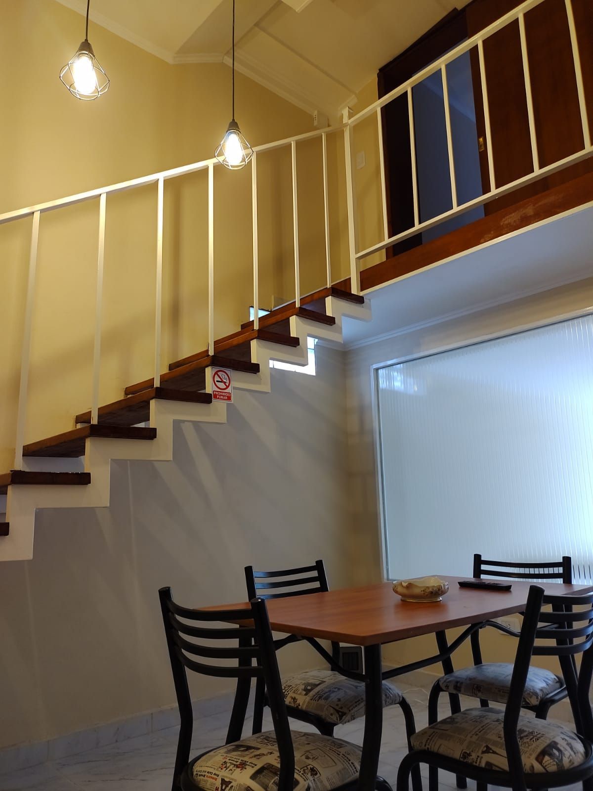 Un comedor con escaleras que conducen a un altillo. Mesa de madera oscura, sillas negras, barandilla blanca y lámparas colgantes.