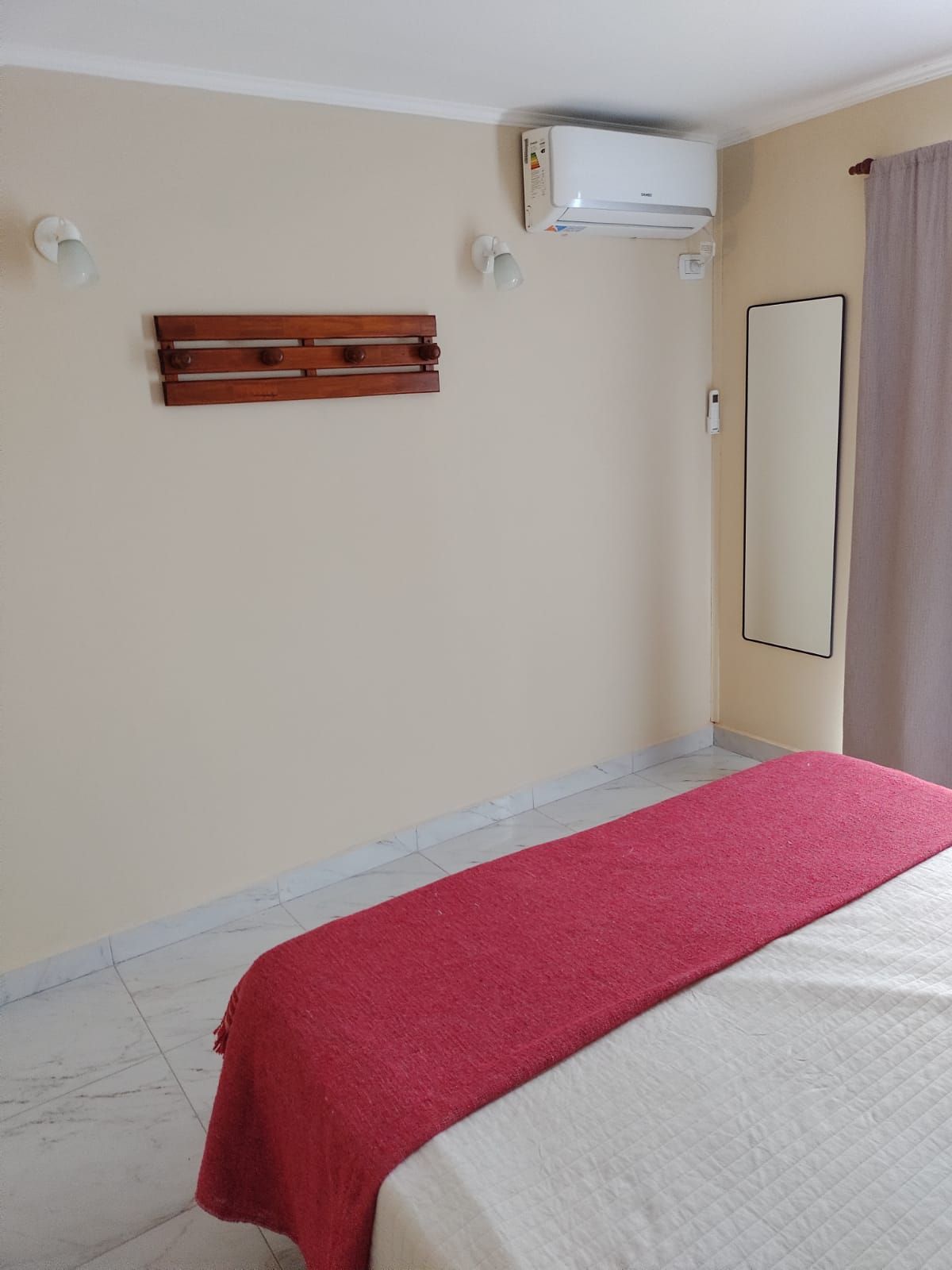 Dormitorio con una cama cubierta con una manta roja, un perchero de madera, un espejo largo y una unidad de aire acondicionado.