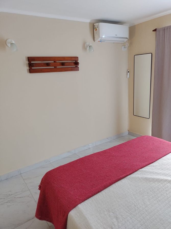 Dormitorio con colcha roja, espejo de cuerpo entero y perchero de madera. Paredes beige claro.