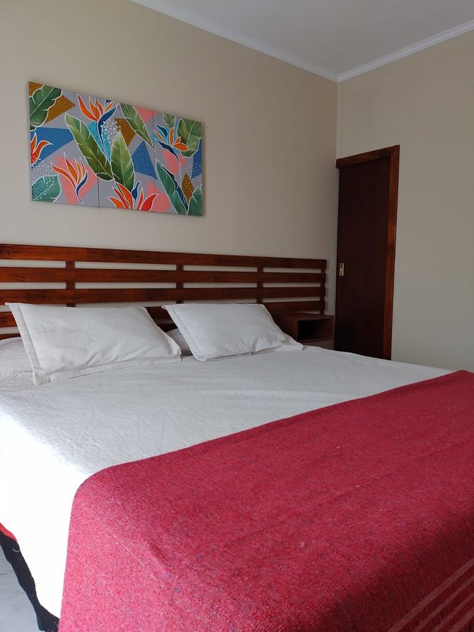 Dormitorio con cama, manta roja, ropa de cama blanca, cabecero de madera, obra de arte floral y una puerta marrón.