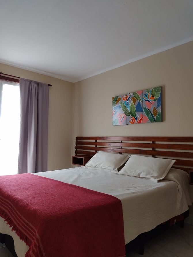 Dormitorio con cama, manta roja, cabecero de madera, cuadro, cortinas, paredes neutras.