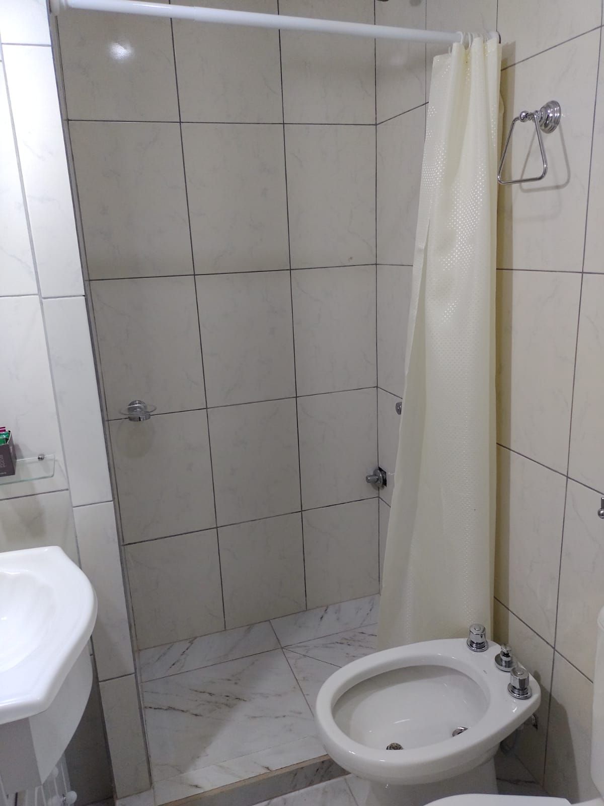 Baño con ducha, inodoro y lavabo. Azulejos blancos en las paredes y cortina de ducha cerrada.