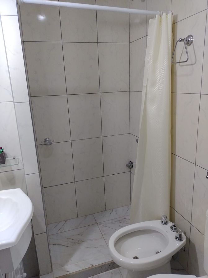 Baño con ducha alicatada, inodoro y lavabo. Cortina de ducha corrida. Paredes blancas y brillantes.