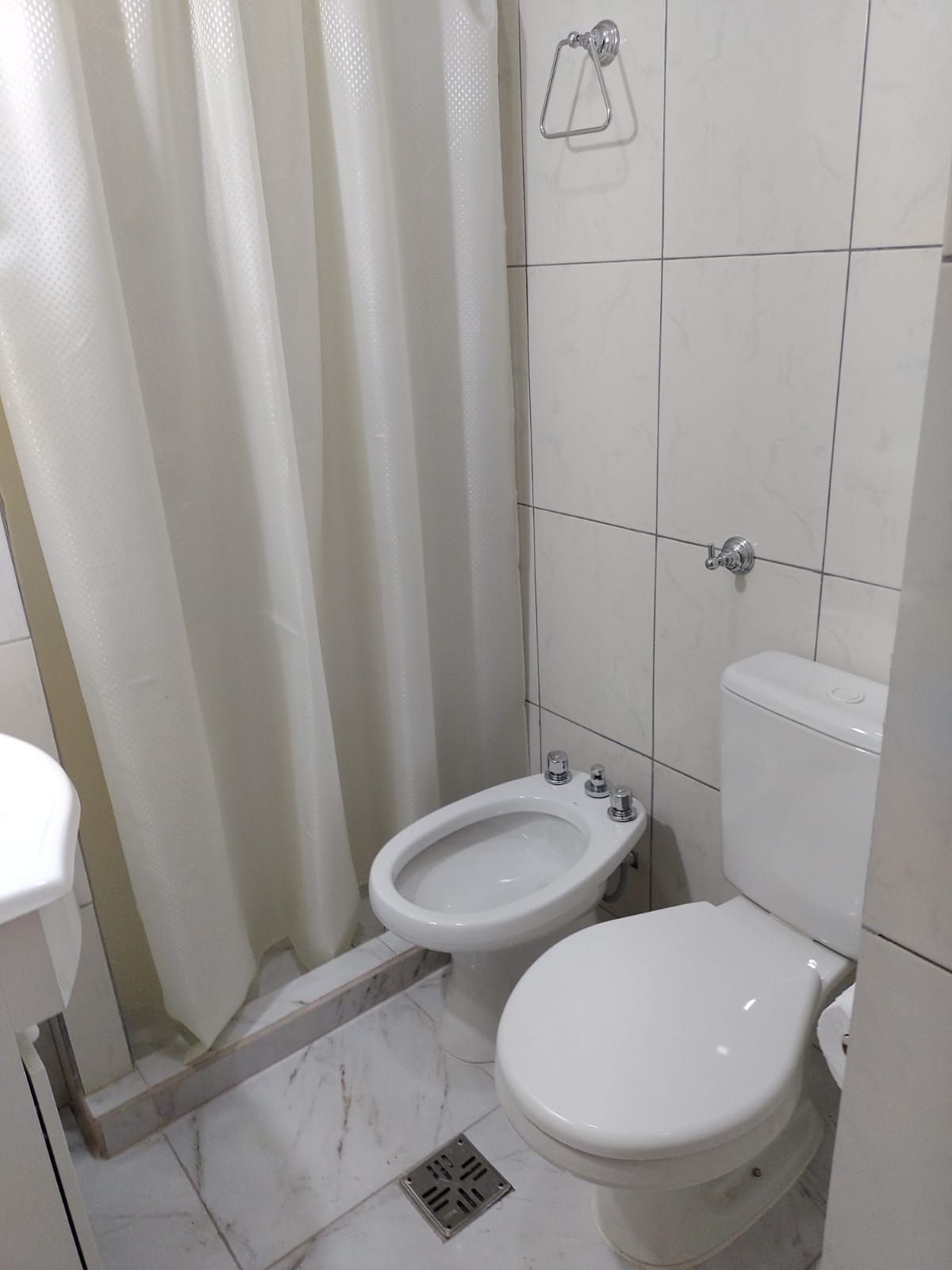 Baño con inodoro blanco, bidet, cortina de ducha y paredes y suelo de azulejos.