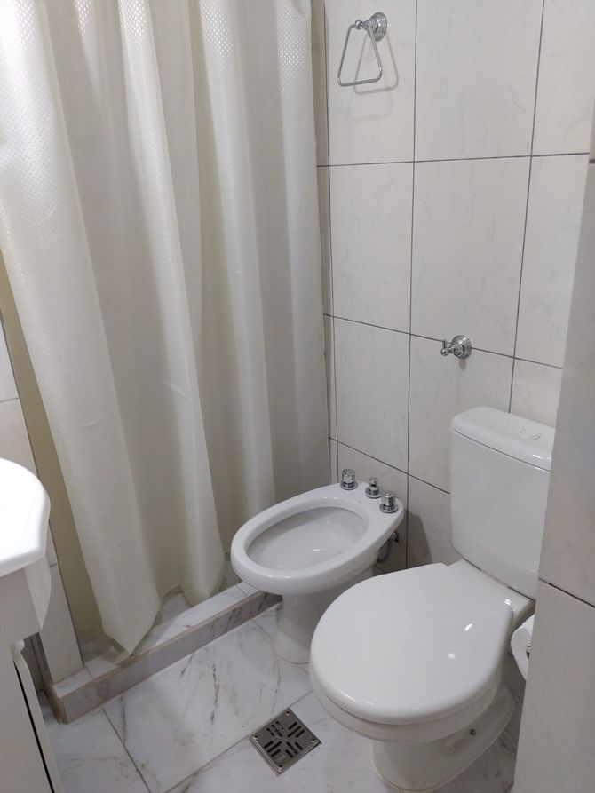 Baño blanco con inodoro, bidé y cortina de ducha. Azulejos y desagüe en el suelo.