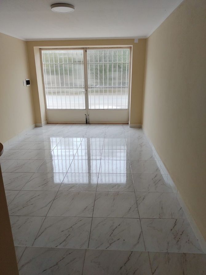 Habitación vacía con paredes de color crema, suelo de baldosas con aspecto de mármol y una puerta de cristal con barras de metal blanco.
