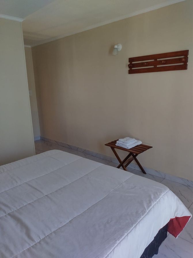 Dormitorio con cama blanca, mesita con toallas y decoración de madera en la pared. Paredes y suelo beige.