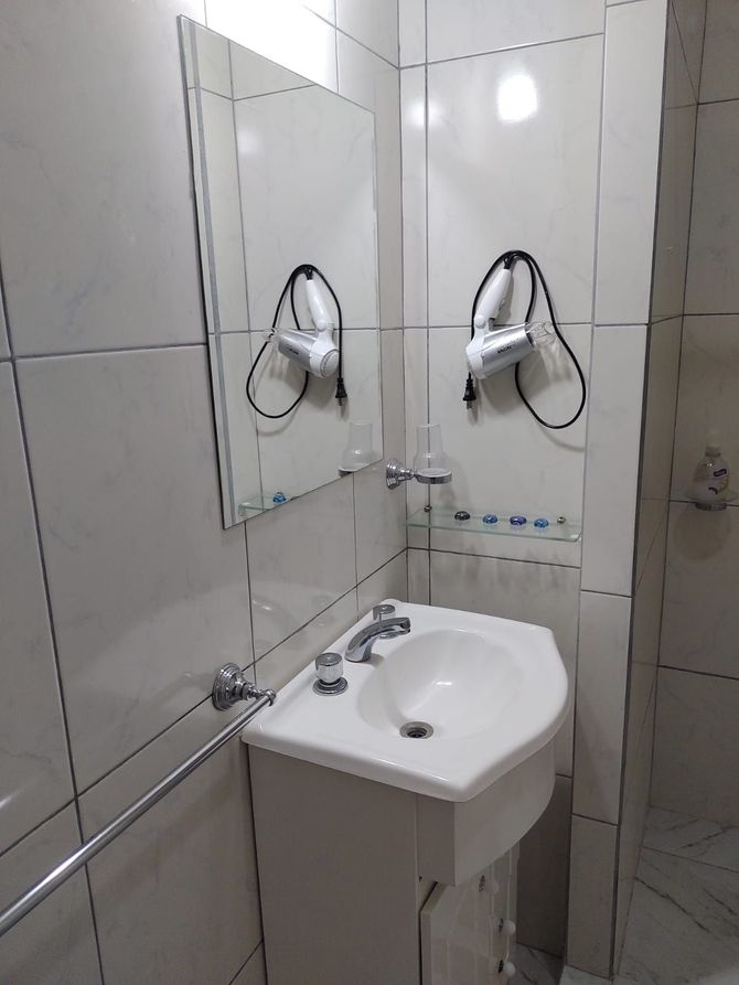 Pequeño baño con azulejos blancos, lavabo, espejo y secador de pelo colgado en la pared.
