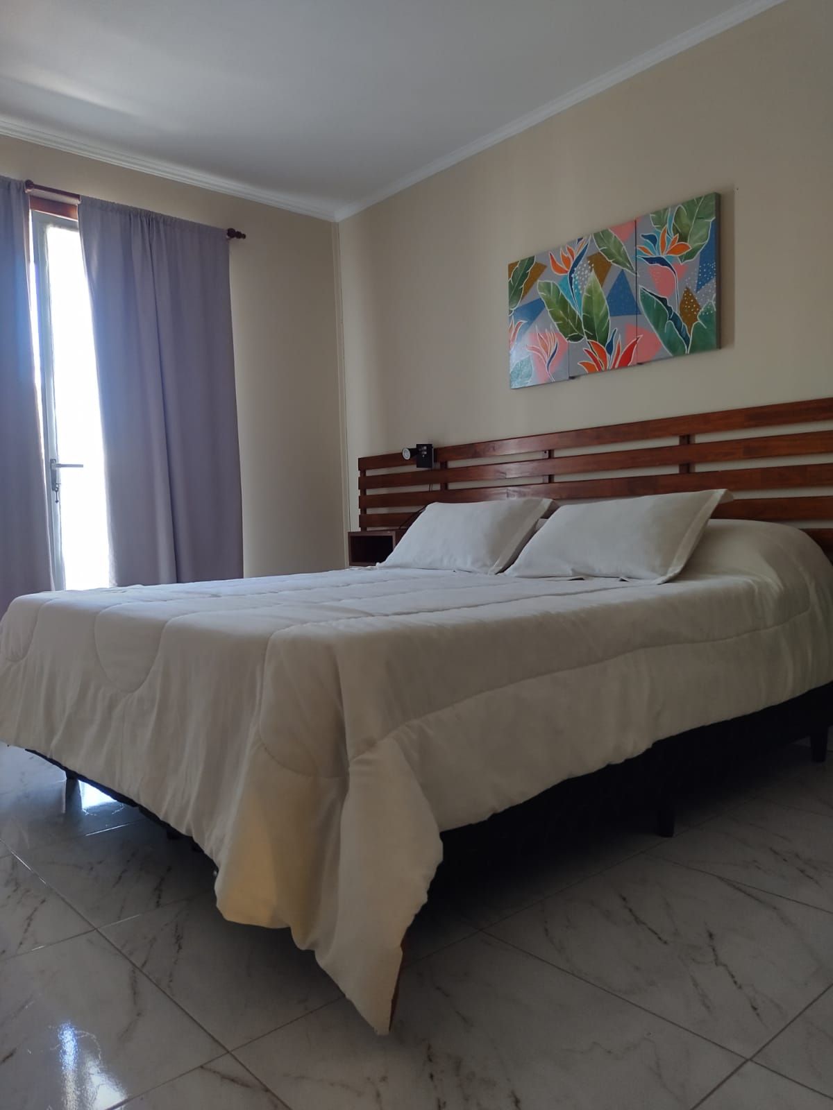 Dormitorio con cama grande, cabecero de madera, obra de arte floral y una ventana con cortinas moradas.