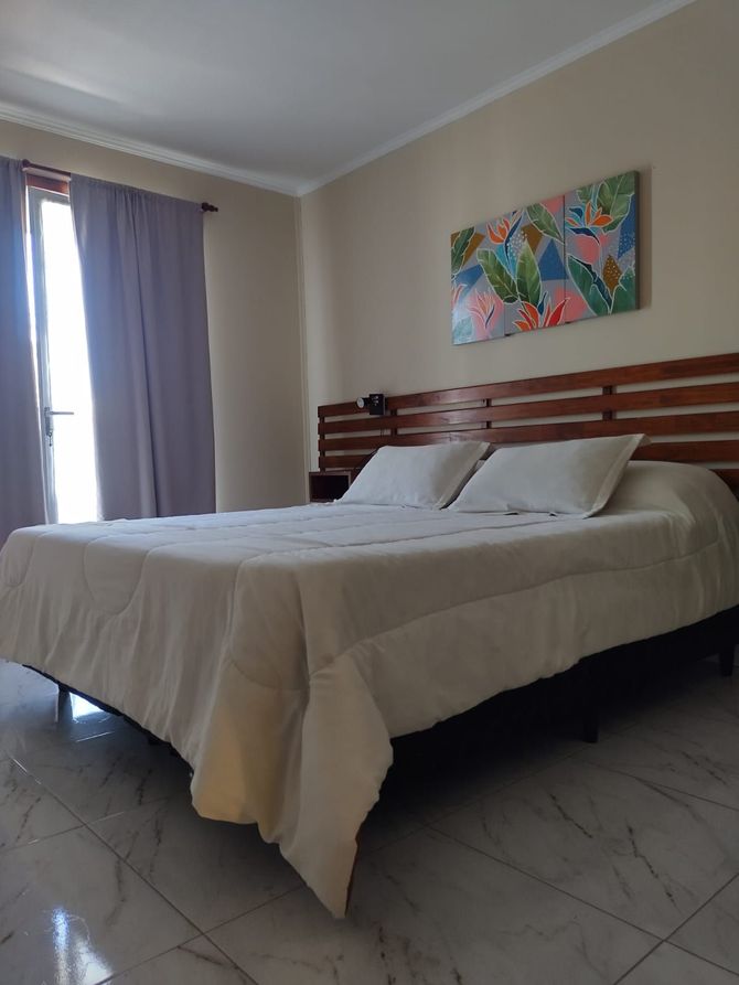 Dormitorio con cama grande, sábanas blancas, cabecero de madera y un cuadro colorido.